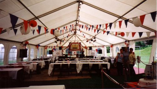Interior of a large event tent decorated with colorful bunting and flags, featuring long tables set for dining. The space is well-lit and has a festive atmosphere, ideal for gatherings or celebrations.