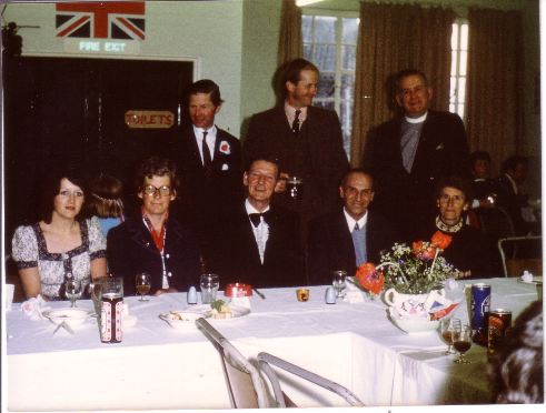 Group of people gathered around a table at a formal event, with men in suits and women in elegant attire, set against a backdrop featuring a British flag. The scene captures a moment of celebration or ceremony, showcasing a mix of smiles and camaraderie.