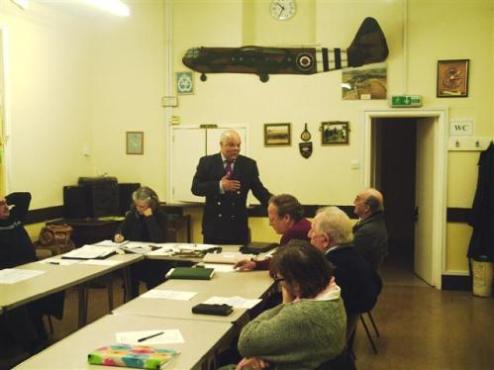A group of people seated around a table in a meeting room, with a speaker standing and addressing the audience. The room features military memorabilia, including a model aircraft hanging on the wall.
