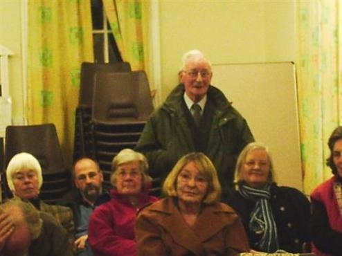 A group of people seated in a community hall. One elderly man stands in a green coat, while others, including women with light and dark hair, sit in chairs with a backdrop of patterned curtains.