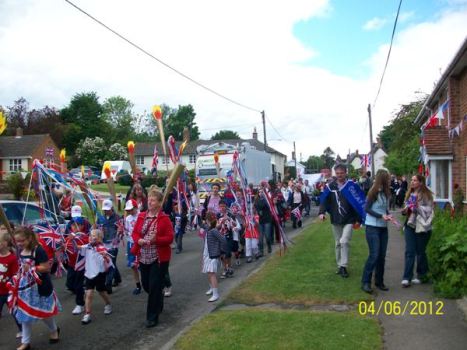 Children and adults participating in a festive parade, carrying flags and banners, walking down a street lined with greenery, under a partly cloudy sky.