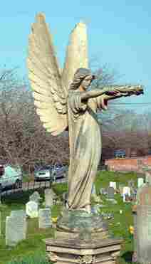 Angel statue with outstretched arms in a cemetery, showcasing intricate details and majestic wings, surrounded by gravestones and trees.