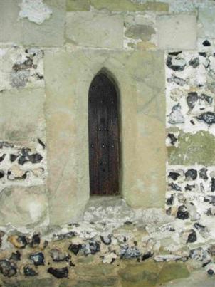 A small, arched wooden door set into a stone wall with a mixture of rough stones and smooth mortar. The door appears old, with a dark finish and decorative metal studs, surrounded by weathered stones.