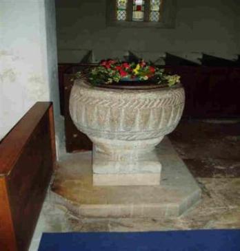 A stone baptismal font with decorative carvings, topped with flowers, located in a church interior. Wooden pews are visible in the background, and a blue carpet is laid on the floor.