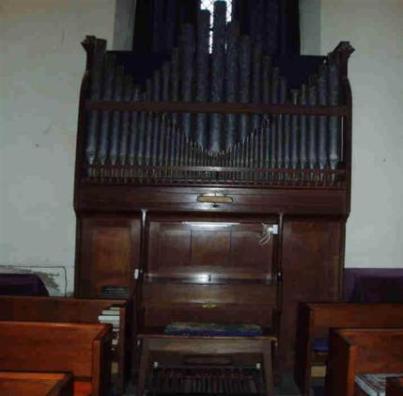 Historic pipe organ in a church interior, showcasing intricate wooden craftsmanship and multiple pipes, with wooden pews visible in the foreground.