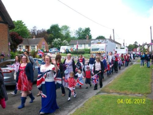 Community parade celebrating a local event, featuring participants of all ages dressed in patriotic attire and waving flags along a rural street.