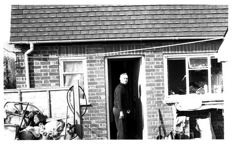 A man is standing at the doorway of a brick house, looking towards the camera. The area around the house is cluttered with various items, including bags and tools.