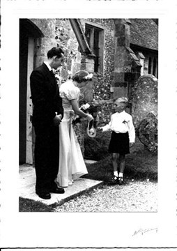 A black and white photograph showing a man in a suit and a woman in a formal dress handing a medal to a young boy in shorts outside a stone building.