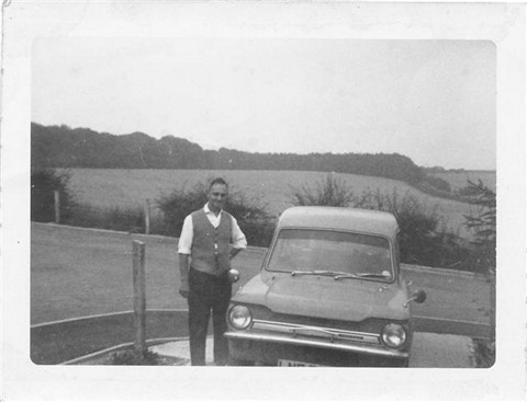A man in a vest stands next to a vintage car in a rural landscape, with hills in the background. The image is in black and white, capturing a nostalgic moment from the past.