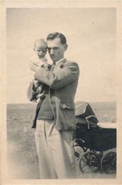 Black and white vintage photograph of a man holding a baby outdoors, with a vintage stroller in the background. The man is dressed in a suit and appears to be smiling at the child, capturing a moment of familial affection.