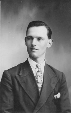 Black and white portrait of a young man in a formal suit and tie, featuring a patterned tie and handkerchief, with a neutral expression and a plain background.