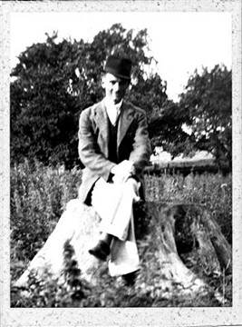 A young man sitting on a log in a field, wearing a suit jacket and light-colored trousers, with trees and a house in the background, captured in black and white.