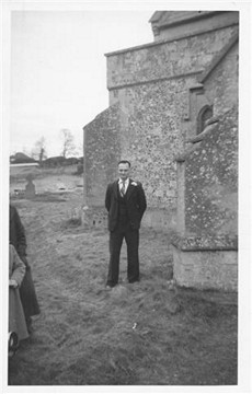 A man in a suit stands near a stone building, surrounded by a grassy area, with trees visible in the background. The image captures a vintage black-and-white photograph.