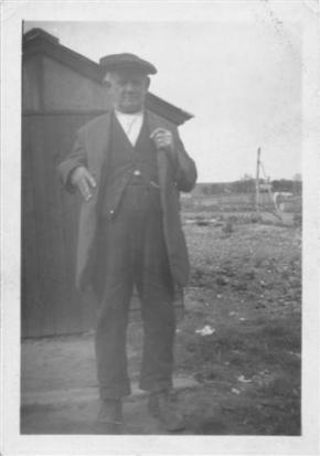 An older man stands outside a small wooden structure, wearing a flat cap, a dark coat, and trousers. He holds a cane in one hand and appears to be posing for the photograph amidst a rural background with sparse scenery.
