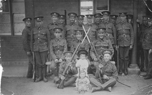 Historical group portrait of soldiers in military uniforms, standing and sitting in formation, with rifles and a child in front, taken in a period setting.