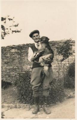A vintage black and white photograph of a smiling man in traditional clothing, holding a small monkey in his arms, with a stone wall and greenery in the background.