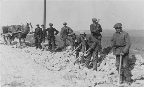 A group of men working on a road construction project, using shovels and pickaxes near a cart drawn by a horse. They are dressed in early 20th-century work clothes, with a rural landscape in the background.