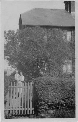 Black and white photograph of a person standing by a wooden fence in front of a house, surrounded by greenery and trees, capturing a nostalgic residential scene.