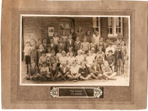 A black-and-white photograph of a large group of school children and teachers, standing and sitting in front of a brick school building. The image has a faded background and a caption at the bottom that reads 'The School TIL-SHEAD', dated 1949.