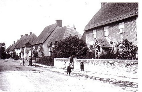 Historic black and white photograph of a quaint village street featuring traditional thatched-roof houses, with people walking along the road and greenery lining the paths, capturing the charm of rural life in the early 20th century.