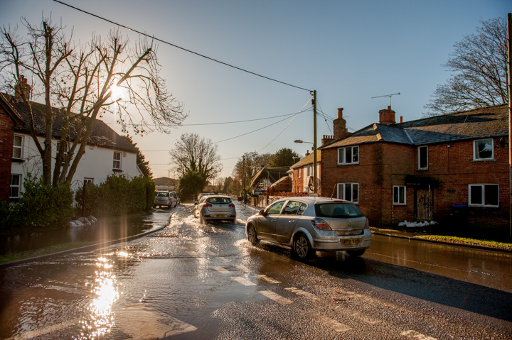 Wet street scene in a residential neighborhood with sunlight reflecting on the pavement, showcasing houses and parked cars.