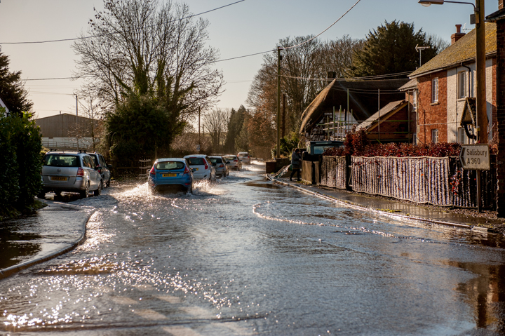 Flooded street with vehicles navigating through water, surrounded by residential homes and trees, illustrating the impact of heavy rainfall.