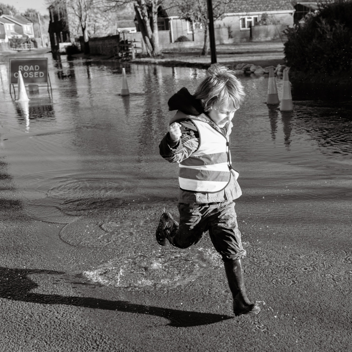 Child in a reflective safety vest joyfully splashing through a puddle on a flooded road, showcasing the playful spirit of childhood in a black and white setting.