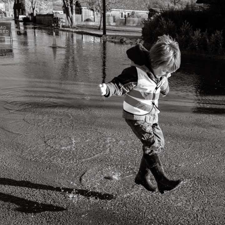 A child in a reflective vest and rubber boots jumping in a puddle on a flooded street, surrounded by trees and buildings, captured in black and white.