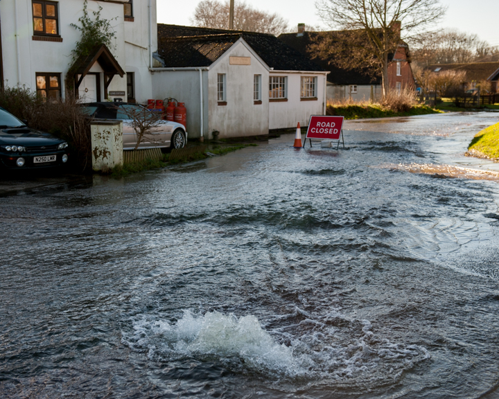 Flooded residential street with water covering the road, obstructing traffic, and warning signs indicating hazards. Nearby houses are partially submerged, illustrating the impact of heavy rainfall or flooding in the area.