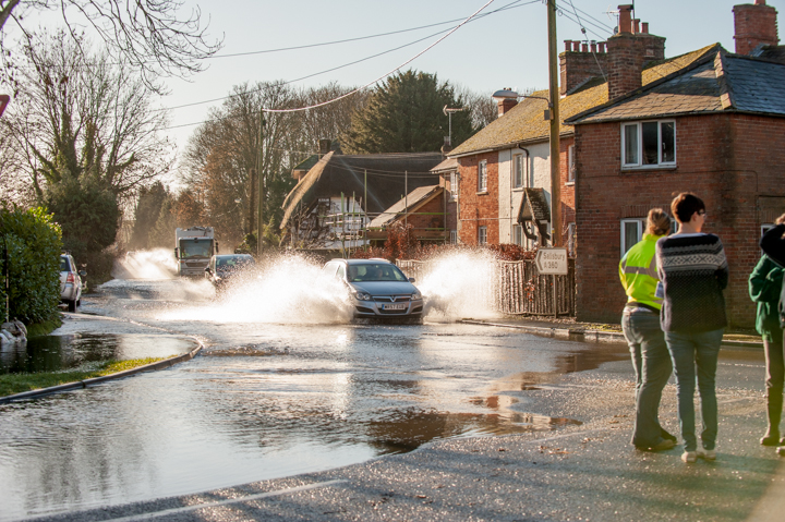 Cars driving through a flooded street, creating splashes, while pedestrians observe from the sidewalk. Nearby houses and trees are visible, indicating a residential area affected by heavy rain or flooding.