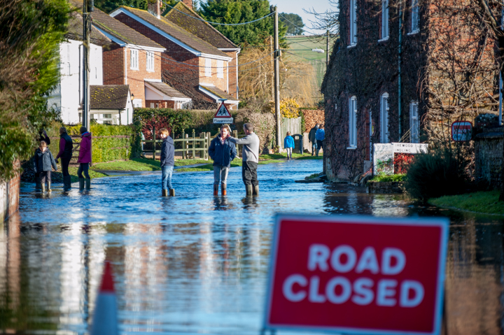 A flooded street with several people walking through shallow water, surrounded by houses and greenery. A road closed sign is prominently displayed in the foreground.