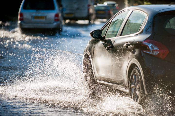 Black car driving through a flooded street, splashing water in the process, with additional vehicles visible in the background.