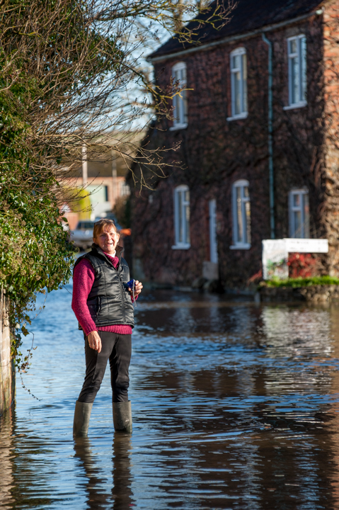 Person standing in floodwaters in front of a brick house, wearing a vest and holding a mobile device, with surrounding greenery and clear skies visible.