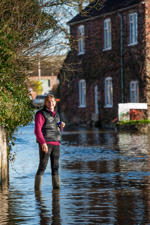 Person standing in floodwaters near a residential area, with partially submerged houses in the background, illustrating the impact of severe flooding.