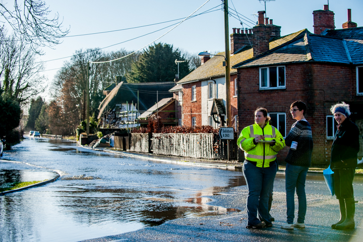 Emergency responders assessing flood damage on a residential street, with homes in the background and standing water on the road.