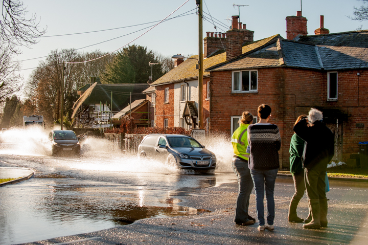 People observing a flooded street with water splashing from passing cars, alongside residential buildings and trees in the background.
