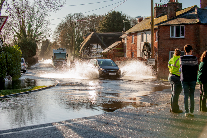A scene of a flooded street with a car driving through the water, creating splashes. People stand on the sidewalk observing the situation, surrounded by residential buildings and trees.
