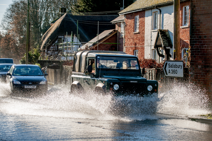 A rugged black SUV splashes through a flooded street in a small village, surrounded by residential buildings and other vehicles on a sunny day.