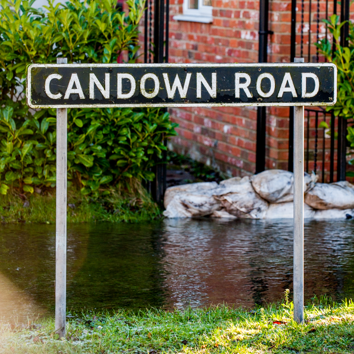 A street sign showing 'Candown Road' positioned beside a small body of water, with greenery in the background and brick buildings nearby.