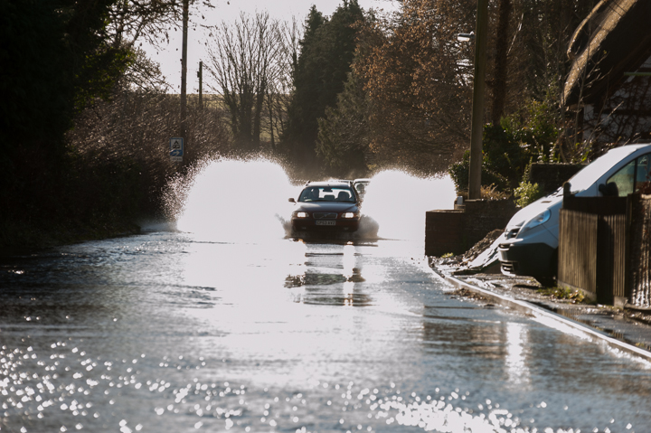 Car driving through a flooded street, creating splashes in the water, with trees and parked vehicles visible in the background.