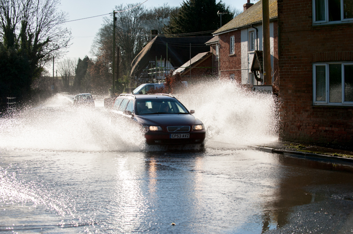 Car driving through flooded street, creating splashes of water, with houses in the background under clear blue sky.