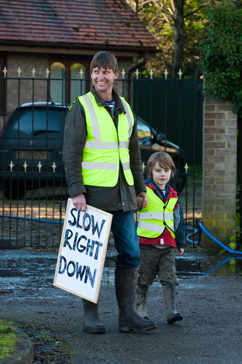 A woman and a young boy wearing bright yellow safety vests stand on a road, holding a sign that says "SLOW RIGHT DOWN" to promote safety. In the background, a residential area is visible, emphasizing the importance of caution in neighborhoods.