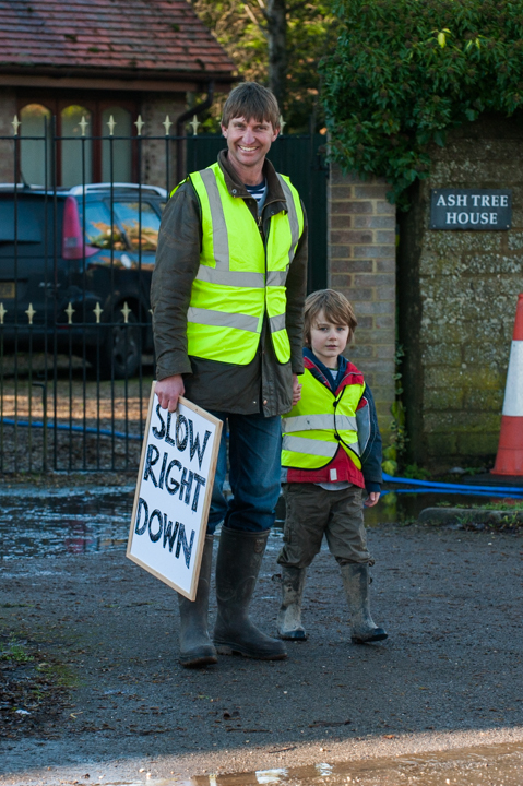 Adult and child wearing high-visibility vests, standing on a road with a sign that reads "Slow Down." They are positioned near a residential area, promoting safety for pedestrians.