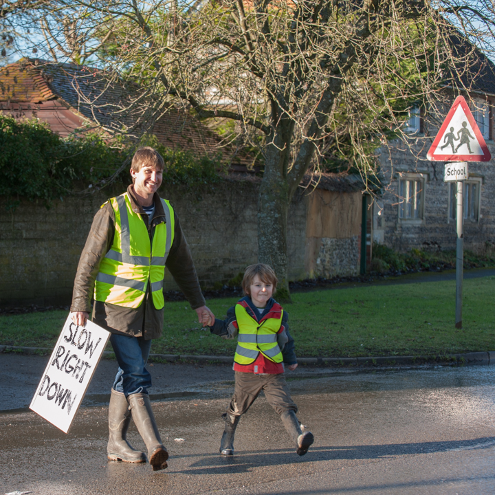A person and a child walking hand in hand on a road, both wearing yellow high-visibility jackets and boots. The adult holds a sign that says 'SLOW RIGHT DOWN.' A school zone sign is visible in the background.