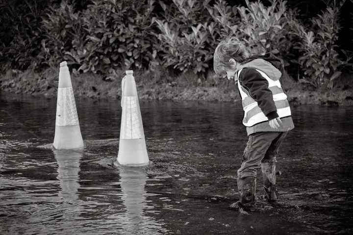 Child in a reflective vest wading through floodwaters, observing two traffic cones in a black and white setting, highlighting the impact of heavy rain or flooding.