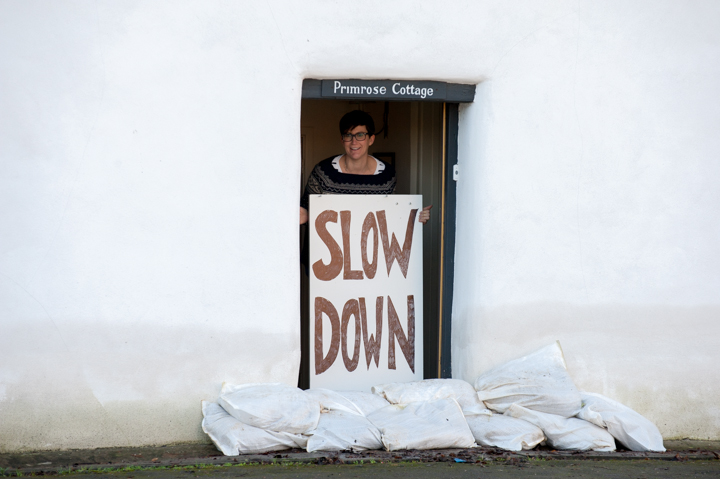 A person standing in the doorway of a cottage holding a large sign that says 'SLOW DOWN.' Sandbags are placed on the ground in front of the entrance.