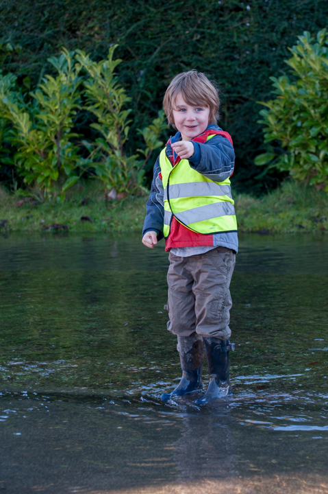 A young boy in a reflective vest and rubber boots standing in shallow water, pointing with a smile. Lush greenery is visible in the background.