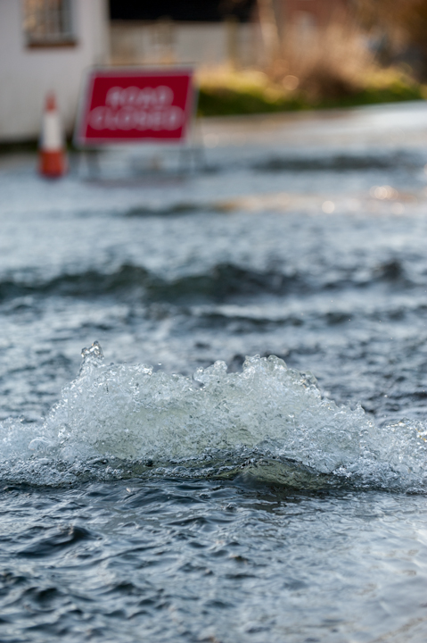 A close-up photo of rippling water in a flooded area, with a red 'Road Closed' sign partially visible in the background, indicating an area affected by flooding.