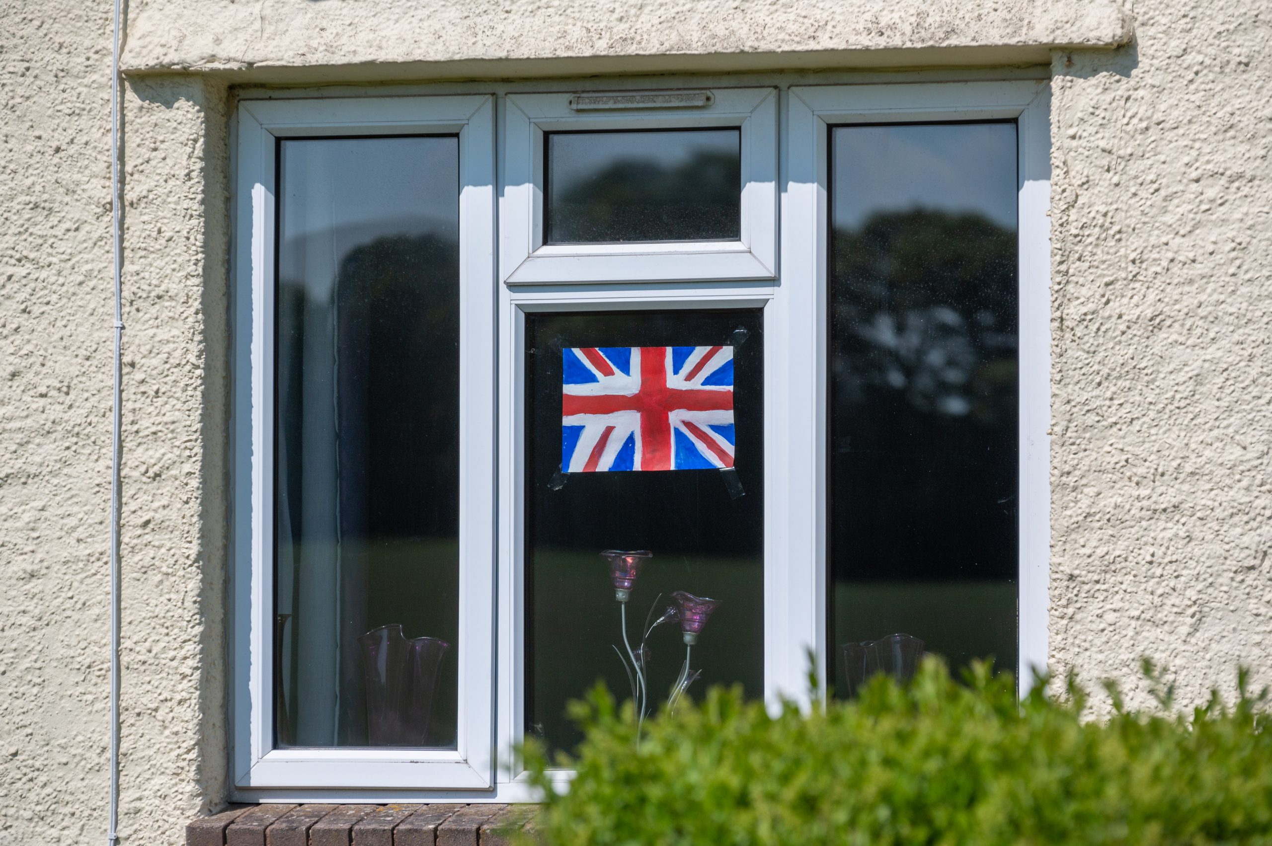 Union Jack flag displayed in a window of a residential building surrounded by greenery on a sunny day.