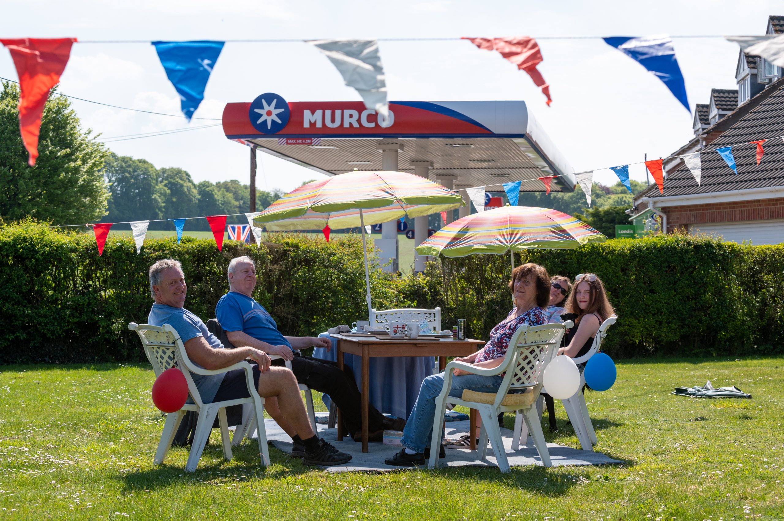 Group of friends enjoying a summer gathering outdoors, sitting around a table with decorations and umbrellas, surrounded by green grass and festive bunting.